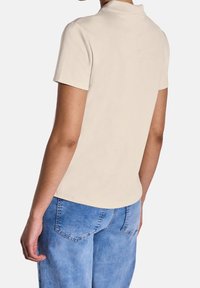 Polo shirt beige à manches courtes avec col, fabriqué en coton, associé à un jean en denim bleu clair, avec des poches arrière.
