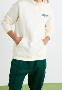 Sweat à capuche de couleur crème avec une poche avant et un cordon de serrage, arborant un logo vert "Converse All-Star". Associé à un pantalon de survêtement vert foncé.
