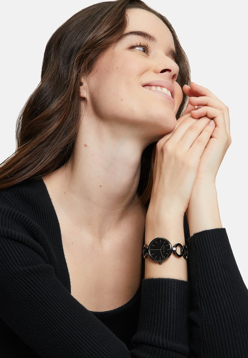 Schwarze Uhr mit rundem Zifferblatt, minimalistisches Design, kombiniert mit einem Gliederarmband. Am Handgelenk getragen, zeigt sie klare Linien und elegante Akzente.