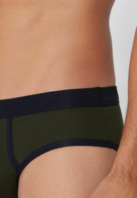 Tmavě zelené boxerské trenýrky s námořnickým pruhem v pase, vyrobené z hladkého materiálu. Mají ploché švy a těsně sedí kolem stehen.