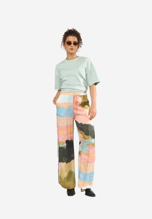 Donna che indossa una maglietta oversize verde chiaro, pantaloni a gamba larga con motivo astratto multicolore, scarpe con tacco nere e occhiali da sole marroni, in piedi con le mani nelle tasche.