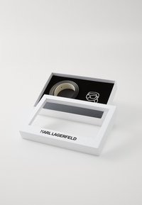 Fehér ajándékdoboz átlátszó fedéllel, amelyben egy fekete és krémszínből készült öv és egy kis logó található. A doboz elején a "KARL LAGERFELD" márkaneve látható.