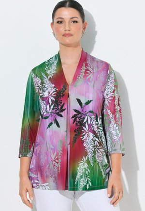 Žena s tmavými vlasy v květovaném, vícebarevném topu kombinujícím zelené, růžové a fialové odstíny, stojící před jednoduchým bílým pozadím.