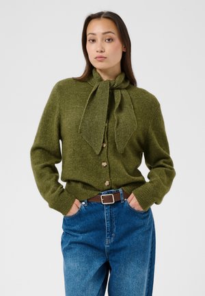 Femme portant un pull boutonné vert olive avec un nœud noué au cou, rentré dans un jean bleu taille haute avec une ceinture marron, les mains dans les poches.