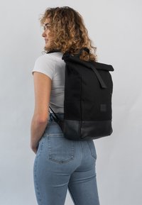 Fekete roll-top hátizsák, textil anyagból készült testtel és bőr aljjal, elülső cipzáras zsebbel és állítható pántokkal. A modell oldalnézetből látható.