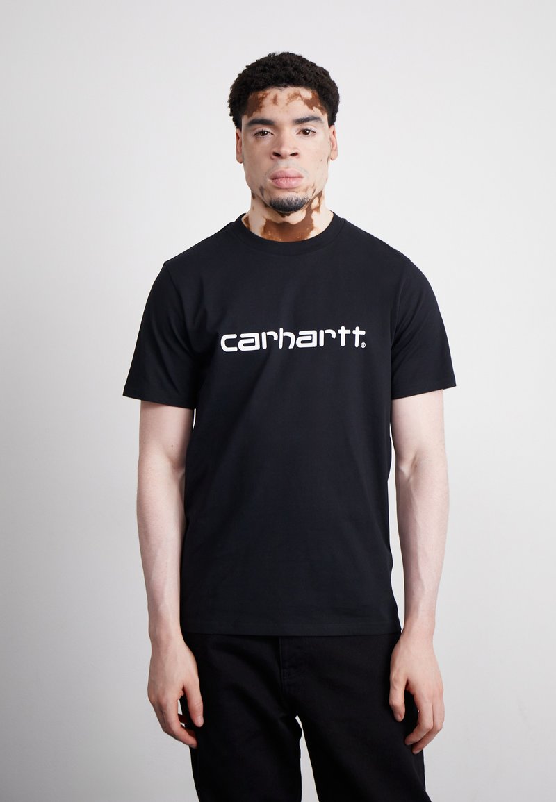 Czarna bawełniana koszulka z białym logo "carhartt." Krótkie rękawy, okrągły dekolt, dopasowany krój. Model stoi z rękami wzdłuż ciała.