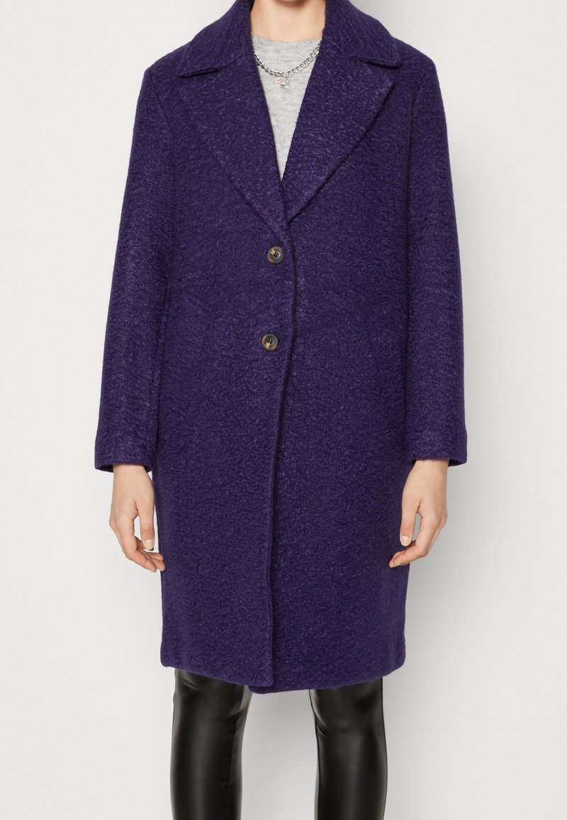 Femme portant un manteau en laine violet foncé à mi-cuisses avec deux boutons, de larges revers, un haut gris, un collier argenté et un pantalon en cuir noir.