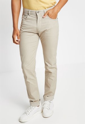 Mann trägt beige schmal geschnittene Hose mit aufgerollten Bündchen, gelbes Hemd ist eingesteckt, dazu weiße Turnschuhe; steht vor einem einfarbigen weißen Hintergrund.