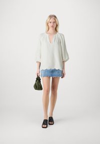 Witte blouse met een V-hals en golvende zoom, voorzien van geborduurde details op de mouwen. Dit is gecombineerd met een denim mini-rok en zwarte slipper-sandalen.