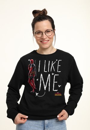 Kvinde med briller og håroppsætning smiler iført sort sweatshirt med Deadpool-figur og teksten "Jeg kan lide mig" på den.