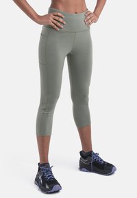 Person iført olivengrønne cropped leggings med sidelomme og sorte og lilla trail runnersko, stående med hænderne på hoften.