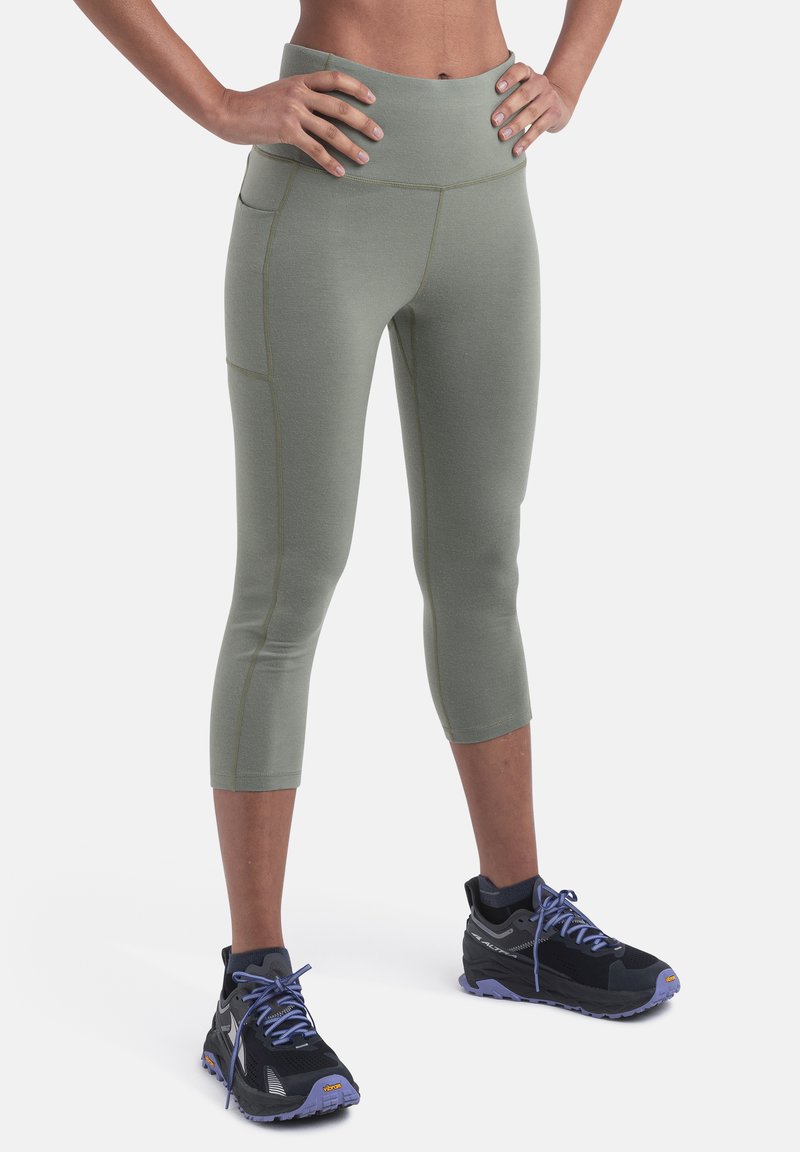 Person iført olivengrønne cropped leggings med sidelomme og sorte og lilla trail runnersko, stående med hænderne på hoften.