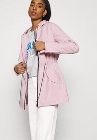 Casaco impermeável rosa claro com a cintura apertada, capuz, fecho com zip frontal e dois bolsos com aba. Usado sobre uma t-shirt gráfica cinza.