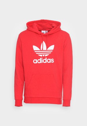 Rode Adidas hoodie met een voorkantzak, een trekkoordcapuchon en een wit Adidas trefoil-logo en tekst op de borst tegen een witte achtergrond.
