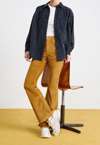 Persona che indossa pantaloni a zampa di elefante in corduroy color senape, sneakers bianche, top bianco a coste e una camicia scura a maniche lunghe, larga, che sta in piedi accanto a una sedia di legno.