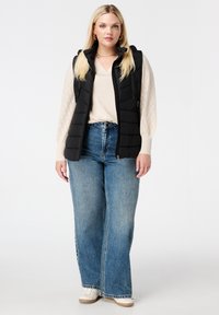 Zwarte gewatteerde vest met ritssluiting, gedragen over een lichtbeige sweater en blauwe wijde jeans. Glad textuur, mouwloos ontwerp, casual look.