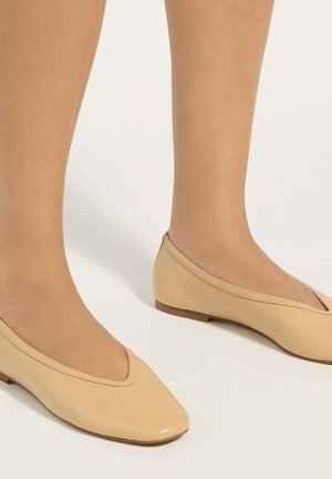 Voeten met beige lage ballerina's met een afgeronde vierkante neus op een witte achtergrond.