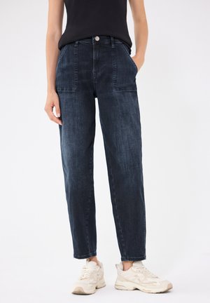Person iført mørkeblå jeans med lige ben og frontlommer med lapper samt hvide sneakers, stående mod en lys baggrund.