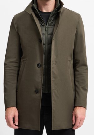 Homme portant un manteau vert olive superposé à une veste matelassée zippée et une chemise noire, debout devant un fond uni.