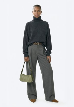 Woman wearing dark gray turtleneck sweater, wide gray trousers, brown pointed shoes, leopard print belt, holding green handbag, standing against white background.