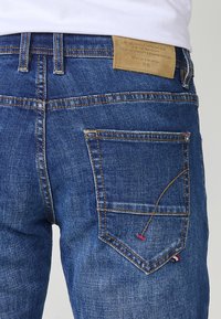 Vaqueros de mezclilla azul con un ajuste relajado, que presentan un diseño de costura amarilla distintivo, un bolsillo trasero con acentos de color y una etiqueta de cuero marrón.