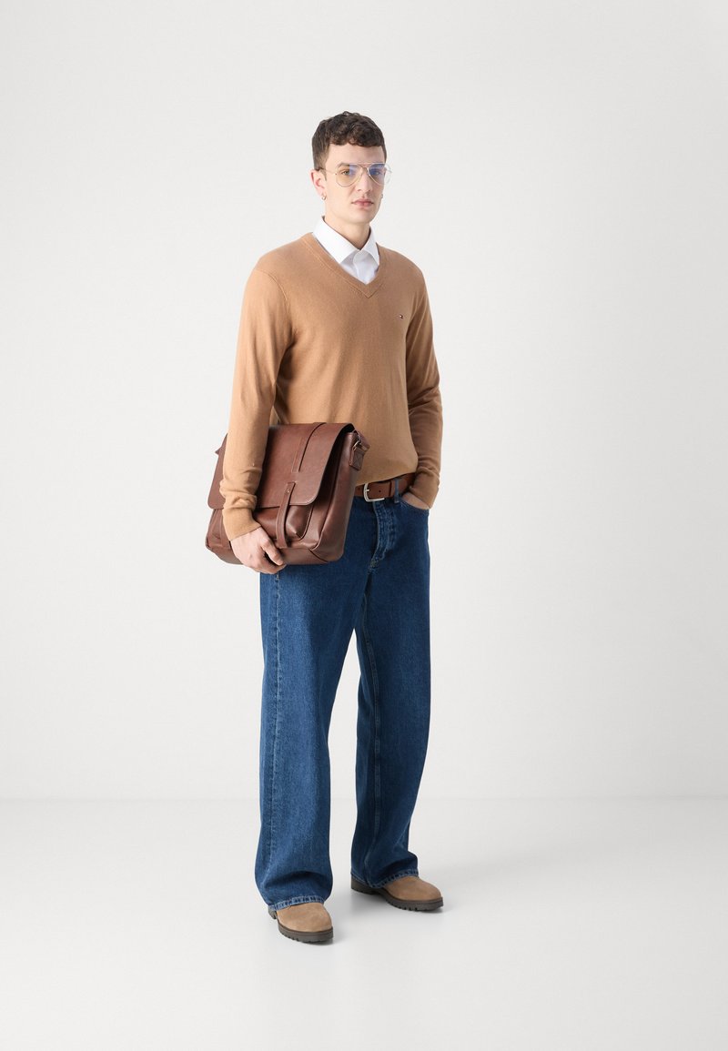 Borsa a tracolla in pelle marrone tenuta da una persona che indossa un maglione beige a scollatura a V, una camicia bianca, jeans larghi blu e scarpe beige.