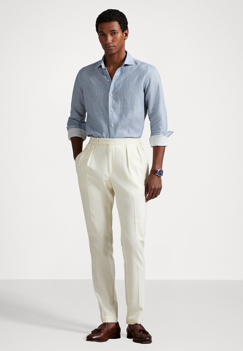 Chemise à motifs bleu clair avec des manches retroussées, pantalon plissé crème et chaussures en cuir marron. Le modèle se tient devant un fond uni.
