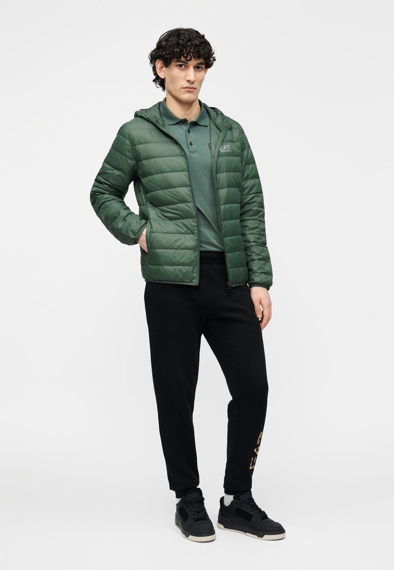 Geacă verde tip puf cu glugă, textură matlasată, asortată cu un tricou polo închis la culoare și pantaloni sport negri, având un detaliu cu logo auriu.