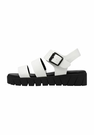 White sandals with three wide straps, featuring a black buckle and a thick, textured black sole. Flat design with a rugged tread pattern.
