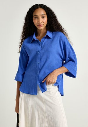Kvinde med langt krøllet hår iført en løs blå skjorte med knapper og en hvid nederdel, stående mod en ensfarvet lys baggrund.