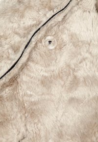 Beżowy materiał zamszowy z miękką, teksturowaną powierzchnią, ozdobiony przezroczystym guzikiem i czarnym szwem wzdłuż krawędzi.