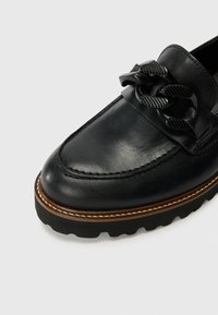 Mocasín de cuero negro con un acento de cadena texturizada, punta redonda y suela de goma gruesa. Presenta costuras en contraste y un acabado suave.