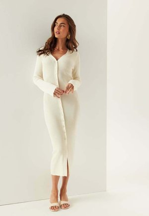 Kvinna i en lång, krämfärgad ribbad klänning med knappar, stående mot en vit vägg, bär beige öppna tå-skor och guldsmycken.