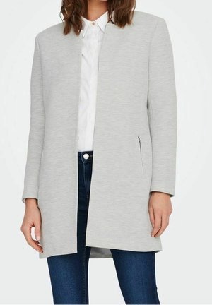 Vrouw die een lichtgrijze blazer met open voorkant draagt over een wit overhemd met knopen en donkerblauwe spijkerbroek.