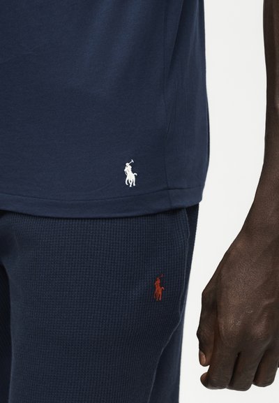 Personne à la peau foncée portant une chemise bleu marine et un pantalon texturé avec de petits logos brodés de cheval et cavalier près de l’ourlet et de la zone de la poche.