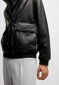 Svart faux skinn bomberjakke med ribbestrikkede mansjetter og liv, som har en stor frontlomme og subtil merkevaredetalj.