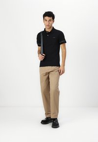 Čierne polo tričko s logom na hrudi a vzorovanými rukávmi, skombinované s khaki nohavicami a čiernymi topánkami, so štýlovým dizajnom a strihom.