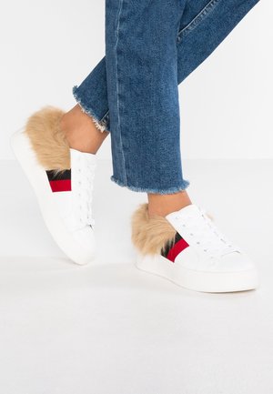 Białe sneakersy z jasnobrązowym futrzanym wykończeniem, czerwono-czarne paski boczne, noszone z niebieskimi dżinsami ze strzępionym zakończeniem na jednolitym białym tle.