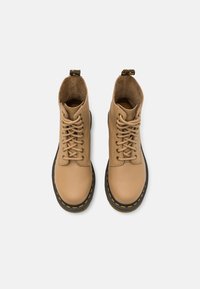 Stivaletti lacci beige in pelle con suole nere e cuciture gialle, visti dall'alto su uno sfondo bianco.