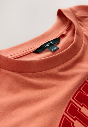 Kortærmet, rundhalset T-shirt i koralorange bomuld med stort rødt grafisk print og sort mærke ved halsudskæringen.