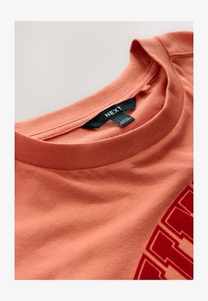 Kortærmet, rundhalset T-shirt i koralorange bomuld med stort rødt grafisk print og sort mærke ved halsudskæringen.