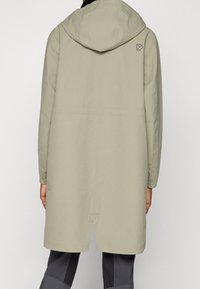 Světle šedý dlouhý kabát s kapucí, vyrobený z hladkého materiálu. Obsahuje decentní logo na zadní straně a elastické manžety. Délka do poloviny stehna.