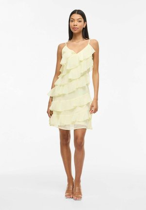 Femme portant une robe jaune pâle sans manches à volants avec bretelles spaghetti et sandales à talons hauts transparentes, debout devant un fond blanc.