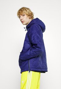 Giacca imbottita viola con cappuccio, cerniera frontale e tasche laterali, indossata sopra pantaloni da jogging gialli vivaci con strisce bianche. Texture liscia.