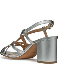 Sølv hælsandal med metalliske stropper, justerbar ankelspenne og en kraftig hæl. Har åpen tå og glatt brun innersåle.