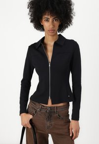 Black, zippered jacket with a collar, structured fit, and long sleeves. Worn with brown pants and a black handbag.