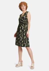 Grünes gemustertes Kleid mit geknoteter Vorderseite, ärmellosem Design und knielangem Saum. Getragen mit beigen Plateau-Sandalen.