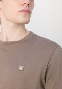 Ruda medvilninė marškinė su apvalia apykakle ir trumpomis rankovėmis. Ant krūtinės yra mažas, tekstūruotas logotipo pleistras. Lygus paviršius ir prigludęs dizainas.