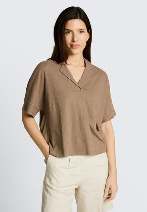 Kobieta z ciemnymi włosami, ubrana w luźną brązową bluzkę z krótkim rękawem i jasnobeżowe spodnie, stojąca z jedną ręką w kieszeni na jednolitym tle.