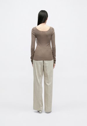 Femme aux longs cheveux foncés, portant un haut marron ajusté à manches longues et un pantalon large beige clair, debout de dos sur fond blanc.